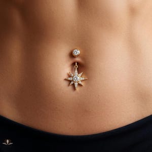 May include: A gold navel piercing featuring a top ball set with a clear stone and a dangling star-shaped charm. The star charm is encrusted with small clear stones and features a larger central clear stone, creating a radiant celestial design.