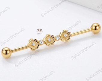 May include: Gold-colored industrial barbell earring featuring three flower designs with clear crystal accents. The barbell has a ball on each end. The jewelry is designed for body piercings.