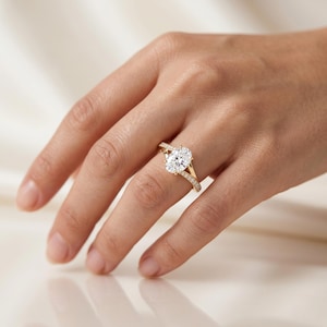 Op de afbeelding: Een gouden ring met een grote ovaal geslepen middensteen die door vier klauwen wordt vastgehouden. De band is verdeeld in twee secties die in het midden samenkomen, waarbij elke kant is ingelegd met kleine, ronde, heldere steentjes.