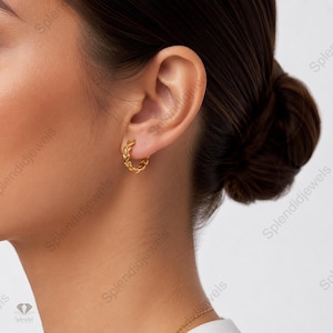 Peut inclure: Gros plan sur une boucle d'oreille dorée en forme de cercle, ornée d'un motif tressé. La personne porte les cheveux foncés attachés en chignon. L'arrière-plan est flou, mettant en valeur le bijou.