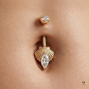Puede incluir: Un piercing para el ombligo de oro con una bola superior con una piedra redonda transparente y un colgante inferior en forma de abanico o concha. El colgante está decorado con una piedra transparente central de talla marquesa.