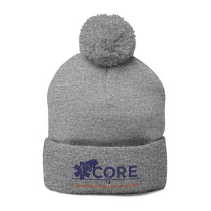 May include: A grey beanie hat with a matching pom-pom. The folded cuff displays the word "CORE" in navy blue, alongside a medical symbol and a heartbeat line. The text "...for the future of healthcare" is below.