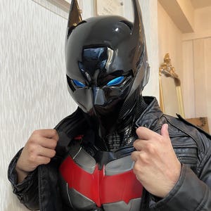Op de afbeelding: Een zwarte Batman-stijl helm met puntige oren en felblauwe ogen. Het borststuk is rood en grijs, en de persoon draagt een zwarte leren jas. Het kostuum heeft een superheldenthema.