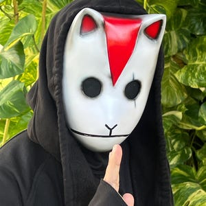 May include: A white mask with black eye holes, red accents, and a black hood. The mask has a cat-like design with red ears and a red stripe down the center. The person is wearing a black hooded sweatshirt.