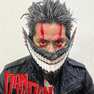 May include: A person wearing a black and white mask with large white teeth and red eyes. The mask is part of a costume, and the person is wearing a black leather jacket. The text "DAN DA DAN" is displayed in red.