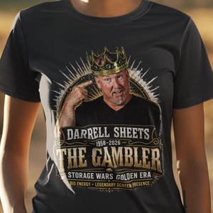May include: Dark gray t-shirt featuring a graphic of a man wearing a crown, with the text "DARRELL SHEETS 1958-2026 THE GAMBLER". Additional text includes "STORAGE WARS GOLDEN ERA BIG ENERGY LEGENDARY SCREEN PRESENCE".