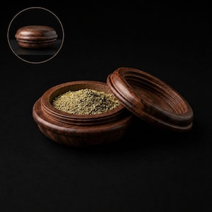Op de afbeelding: Een ronde, donkerbruine houten container met een bijpassend deksel, zowel open als gesloten afgebeeld. De open container is gevuld met een kleine hoeveelheid gedroogde groene kruiden. Het hout heeft een zichtbare nerf en een gladde afwerking.
