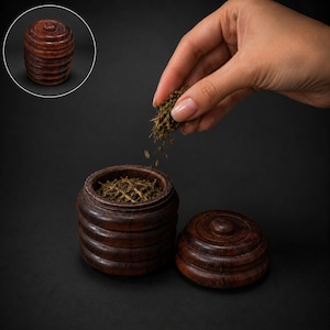 Puede incluir: Un recipiente cilíndrico de madera de color marrón oscuro con detalles acanalados horizontales. El recipiente cuenta con una tapa extraíble a juego con un pequeño pomo. Se espolvorean hierbas secas en el recipiente abierto, que se muestra tanto abierto como cerrado.