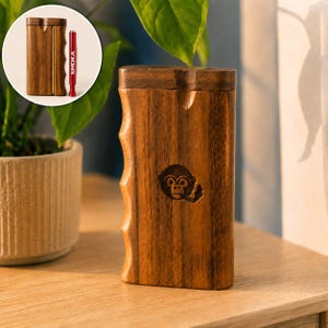 May include: A rectangular wooden dugout box with a natural grain finish and a carved monkey face icon on the front. The side features ergonomic finger grooves. An inset circular image shows the box open with a red metal one-hitter pipe labeled SMOKA.