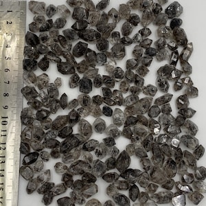 Könnte beinhalten: Eine Ansammlung kleiner, durchscheinender, dunkelgrauer und schwarzer Rauchquarzkristalle mit spitzen Enden, angeordnet neben einem Metalllineal mit Zentimeter-Skala, wobei die Kristalle eine Größe von 0,2 bis 1,5 Zentimetern aufweisen.