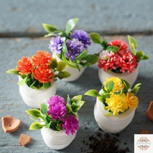 May include: Four small white ceramic pots each containing a cluster of artificial flowers in vibrant colours including orange, purple, red, and yellow. Each plant features green leaves and textured blossoms. A small logo is visible in the bottom right corner.
