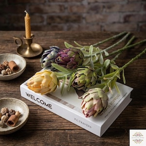 May include: A collection of five artificial artichoke stems in shades of yellow, purple, and green, resting on a white book titled WELCOME and P.L. TRAVERS. The stems feature layered, petal-like bracts and long green stalks with small leaves.