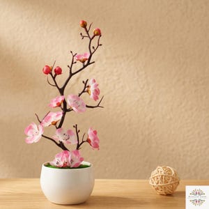 May include: A decorative artificial cherry blossom branch with pink flowers and red buds, arranged in a small, round, white ceramic pot. The plant features dark brown, slender, branching stems and is accompanied by a small, round, woven rattan ball.
