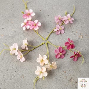 Peut inclure: Cinq tiges de fleurs artificielles disposées en étoile, avec des pétales dans des tons rose clair, magenta et crème. Chaque tige possède des branches en fil vert et des fleurs à cinq pétales. Un petit logo avec le texte Petals by Aisha est visible.