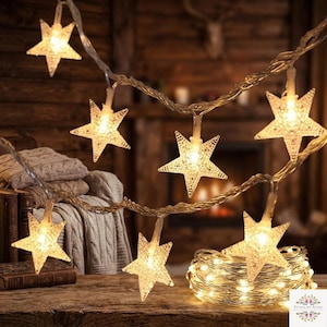 Puede incluir: Una cadena de luces LED con cubiertas transparentes y texturizadas en forma de estrella. Las luces emiten un brillo cálido, con algunas estrellas colgando en línea y otras enrolladas en un paquete sobre una superficie de madera con cable fino.