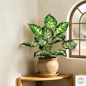 May include: A potted artificial Dieffenbachia plant with large, variegated leaves featuring deep green edges and bright yellow-green centres. The plant is housed in a round, textured, light brown ceramic pot with a wide rim and a matching base tray.