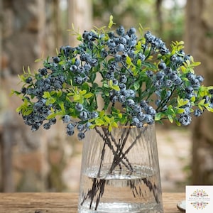 May include: A clear, vertically ribbed glass vase holds a cluster of artificial blueberry stems. The stems feature numerous small, dusty blue berries and bright green leaves, creating a dense, rounded arrangement with visible brown stalks.