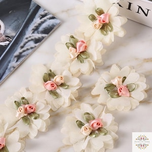 May include: A decorative trim featuring a series of off-white fabric flowers, each accented with two small pink ribbon roses and green ribbon leaves. The floral elements are arranged in a line, with additional individual flowers placed nearby.