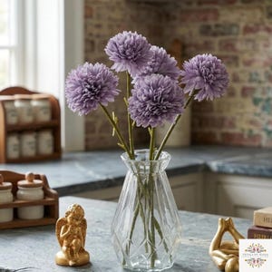 May include: A clear glass vase with a geometric faceted design holds five artificial lavender-colored chrysanthemum flowers on green stems. The arrangement is displayed alongside a small gold-colored angel figurine and a gold-colored abstract sculpture.