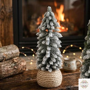 May include: A miniature artificial Christmas tree with flocked, snow-dusted green needles, standing in a round base wrapped in thick, natural-coloured jute rope. The tree has a conical shape and a dense, textured appearance.