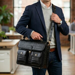 Könnte beinhalten: Schwarze Lederaktentasche mit Krokodilhautstruktur, mit zwei Vordertaschen und einem Schulterriemen. Die Aktentasche hat goldfarbene Beschläge und einen oberen Griff. Die Person trägt einen marineblauen Blazer und Jeans.