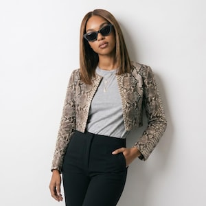 May include: A cropped snakeskin-print jacket with a neutral color palette, paired with a gray t-shirt and black high-waisted pants. The model is wearing black sunglasses and a delicate necklace.