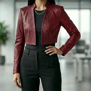 May include: A burgundy leather bolero jacket with long sleeves and intricate floral embossed detailing on the front panels. The jacket is cropped, and the model is wearing a black top and black pants. A gold necklace is visible.