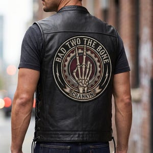 Puede incluir: Chaleco de cuero negro con un gran parche circular en la espalda. El parche presenta una mano de esqueleto haciendo un gesto de rock and roll, con las palabras "BAD TWO THE BONE" y "CRANKIN" en un diseño circular.