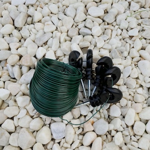 Puede incluir: Un conjunto de aisladores negros para cercas eléctricas con tornillos, una bobina de alambre verde y otros componentes. Los elementos se disponen sobre guijarros blancos, para cercas o proyectos de jardín.