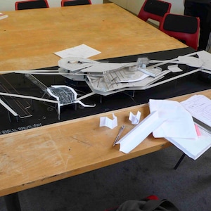Op de afbeelding: Een gedetailleerd architectonisch model, gemaakt van witte en grijze materialen, staat op een zwarte basis. Het model staat op een grote houten tafel, met documenten en gereedschap. De tekst op de basis luidt "SPATIAL SHAPE VERTICAL BRANCH STUDY".