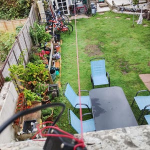 以下が含まれることがあります： 小屋、自転車、庭の家具がある庭の上からの眺め。庭には緑の芝生、木の柵、鉢植えの植物があります。赤いロープが画像の上部から垂れ下がっています。