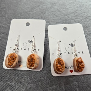 May include: Handmade earrings with a passion fruit design. The earrings are made of clay and have silver-coloured hooks. The passion fruit design is brown and tan with a textured surface. The earrings are displayed on white cards with the word "CLAYATIVE".