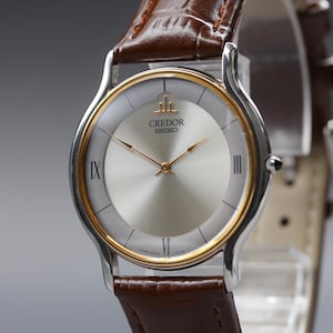 May include: A Credor Seiko wristwatch featuring a round silver dial with gold-tone hands and markers, Roman numerals at the three, six, and nine o'clock positions, a gold-tone bezel, and a brown leather strap with a textured crocodile-like pattern.