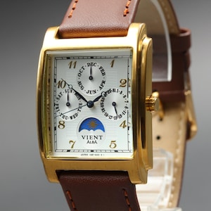 May include: A rectangular gold-tone watch with a white textured dial, brown leather strap, and gold Arabic numerals. The face features three sub-dials for month, day, and date, a moon phase indicator, and the text VIENT ALBA printed in black.