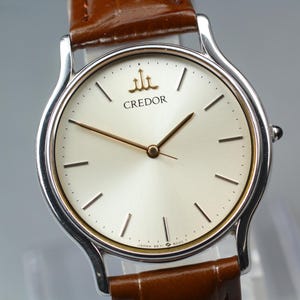 May include: A Credor wristwatch featuring a round silver-tone case, a cream-colored dial with thin gold-tone hour markers, and gold-tone hands. The watch includes a brown leather strap with a textured finish and a small crown on the right side.
