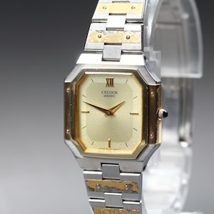May include: A two-tone Credor Seiko wristwatch featuring a gold-colored octagonal dial with a Roman numeral at the twelve o'clock position. The watch has a silver and gold-tone metal link bracelet with visible screw details on the links.