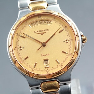 May include: A two-tone Longines quartz wristwatch featuring a gold-colored dial with a day-of-the-week window at the twelve o'clock position and a date window at the six o'clock position. The watch has a textured gold bezel and a metal link band.