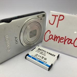 Peut inclure: Un appareil photo numérique Canon IXY argenté avec un objectif zoom, une batterie et un panneau avec l'inscription "JP Camera Can" en rouge. La batterie est blanche avec des accents bleus.
