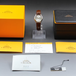 May include: A Credor wristwatch with a silver dial and brown leather strap, displayed with a yellow outer box, a black inner box, two yellow booklets, a white warranty card, a white instruction manual, and a small black product hang tag.