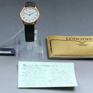 May include: A gold-tone wristwatch with a white dial, black Roman numerals, and a black leather strap. Included are a blue tag with the text "LONGINES", a gold-colored booklet labeled "LONGINES THE LONGINES STYLE", and a light green document with Japanese text.