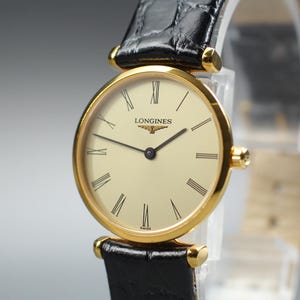 May include: A Longines wristwatch with a round gold-tone case and a cream-coloured dial featuring black Roman numeral hour markers. The watch has thin black hands and is attached to a black leather strap with a textured, embossed crocodile pattern.