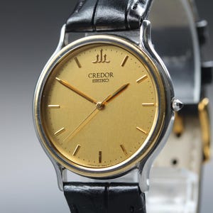 May include: A Credor Seiko wristwatch featuring a round gold-tone dial with thin gold-tone hour markers and hands. The watch has a silver-tone metal case and a black leather strap with a textured finish. The Credor logo is visible at the top.