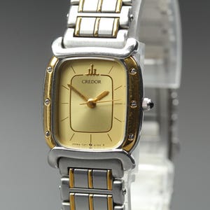 May include: A Credor wristwatch with a rectangular gold-toned dial, gold hands, and a two-tone silver and gold metal link bracelet. The watch face features the Credor logo at the top, thin hour markers, and the text JAPAN 1271-0100 R at the bottom.