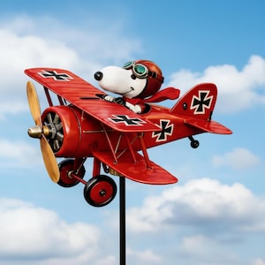 Puede incluir: Un molinillo de viento de avión biplano rojo con Snoopy como piloto, con una bufanda roja y gafas de aviador. El avión tiene cruces negras en las alas y la cola, con una hélice de madera. Fondo de cielo azul nublado.
