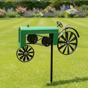 Puede incluir: Estaca de jardín con forma de tractor de metal verde con ruedas y engranajes negros. El tractor decorativo tiene un volante y está colocado en un césped con un jardín de flores al fondo.