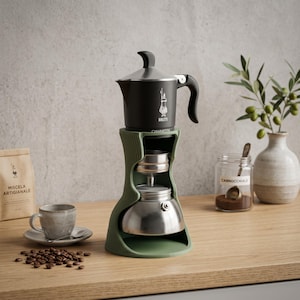 Può includere: Una caffettiera moka Bialetti nera è posizionata su un supporto verticale a coste color verde salvia. La caffettiera presenta una base in argento metallizzato e una camera superiore nera con il logo Bialetti stampato in bianco sul fronte.