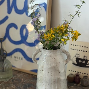 Peut inclure: Un vase en céramique doté de deux anses latérales, présentant un émail blanc cassé moucheté et une base en terre cuite visible. Le vase contient un bouquet de petites fleurs violettes en cloche et de fleurs jaunes en étoile avec des tiges vertes.
