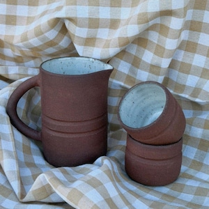 Peut inclure: Une carafe en céramique brun foncé avec une anse incurvée et un intérieur blanc cassé moucheté, accompagnée de deux tasses en céramique assorties empilées, présentant la même finition extérieure brun foncé et intérieure mouchetée.