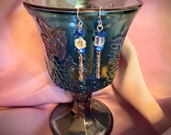 May include: A pair of dangle earrings featuring silver-toned hooks, small blue petal-shaped beads, clear iridescent cube-shaped beads, and metallic silver-toned cylindrical spacers hanging vertically from a dark blue glass goblet.