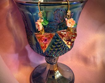 May include: A pair of dangle earrings featuring triangular resin dice with embedded floral elements and red numbering. Each earring is topped with a small pink flower and green leaf charm, suspended from gold-toned metal hooks.